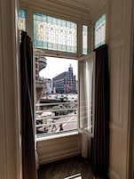 A room with a large window overlooking a busy street, featuring a view of a building with a clock tower.