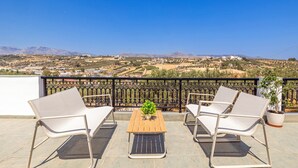 Una terraza con mesa de madera y dos sillas blancas, una planta en maceta y vistas a un paisaje montañoso.