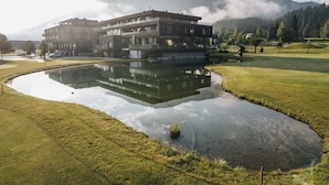 A modern building with a reflection in a pond, surrounded by a golf course and mountains. - Luxurious Penthouse on the Golf Course (Westendorf)