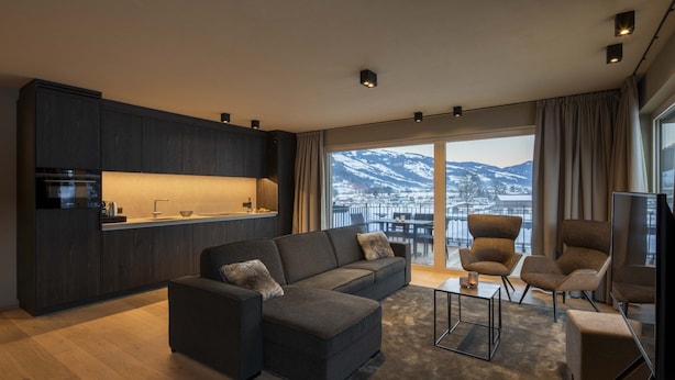A modern living room with a dark-colored sofa, a coffee table, and a TV. There is a kitchen area with dark cabinets and a window overlooking a snowy mountain. - Luxurious Penthouse on the Golf Course (Westendorf)