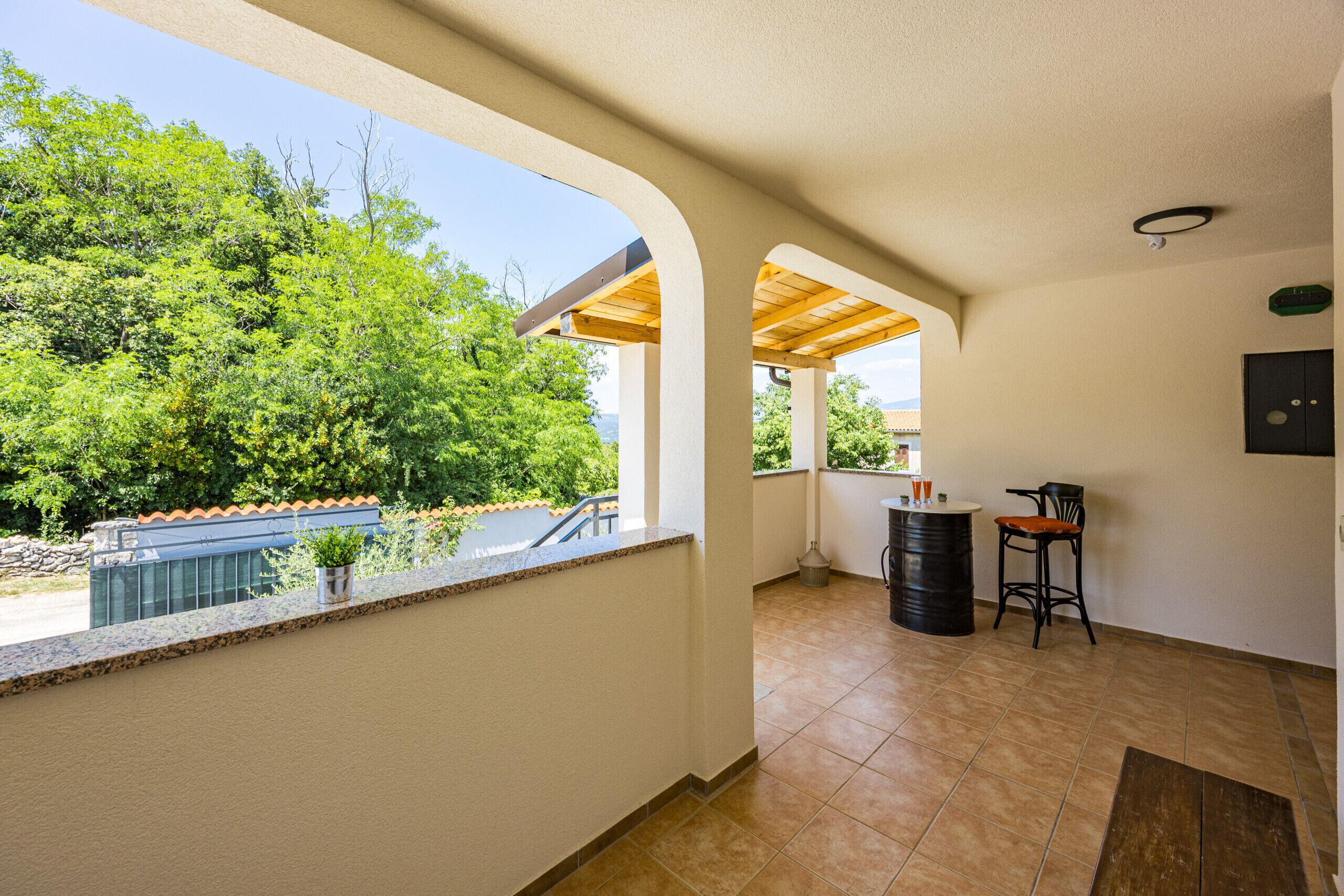 A covered balcony with a view of greenery and a building. There is a small table and two chairs.