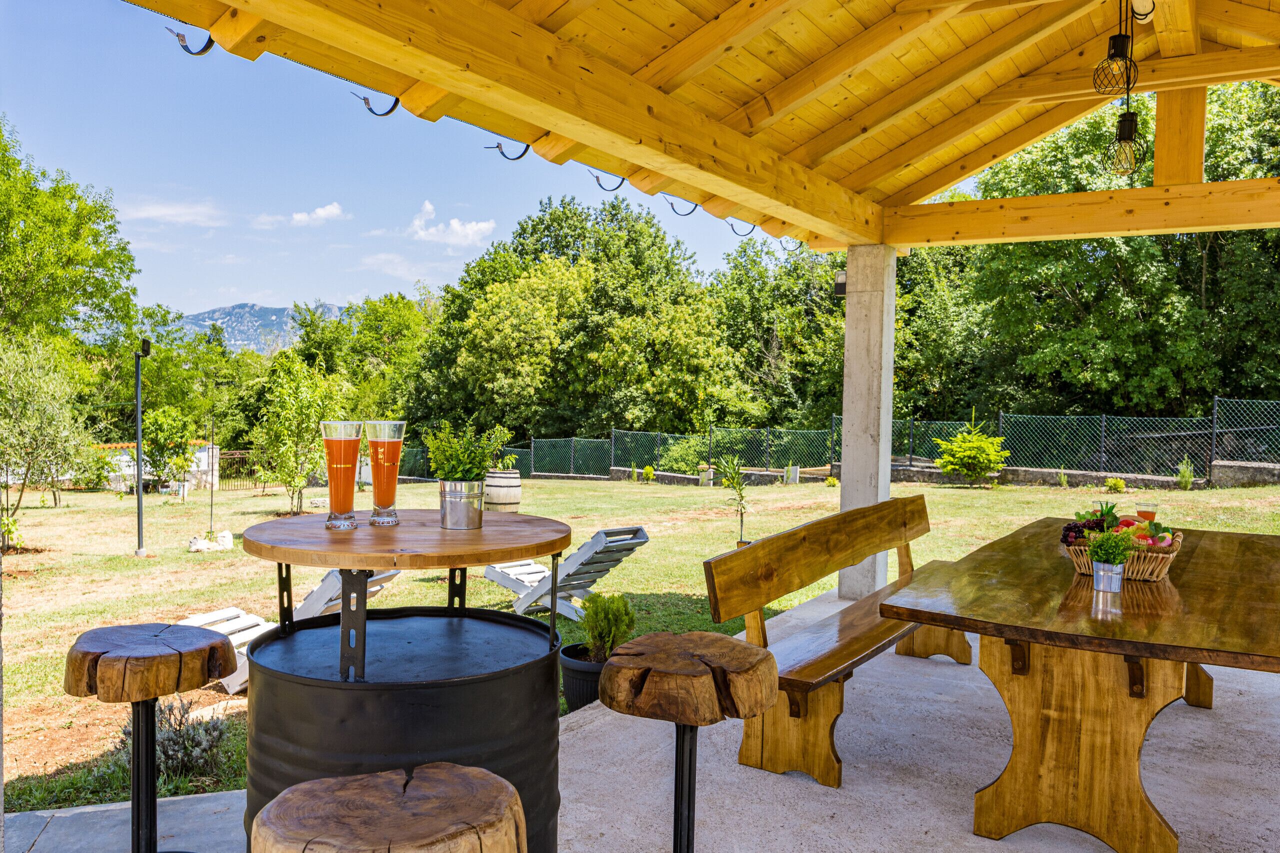 Een terras buiten met een houten tafel en bankjes, omgeven door groen en met uitzicht op bomen en heuvels.