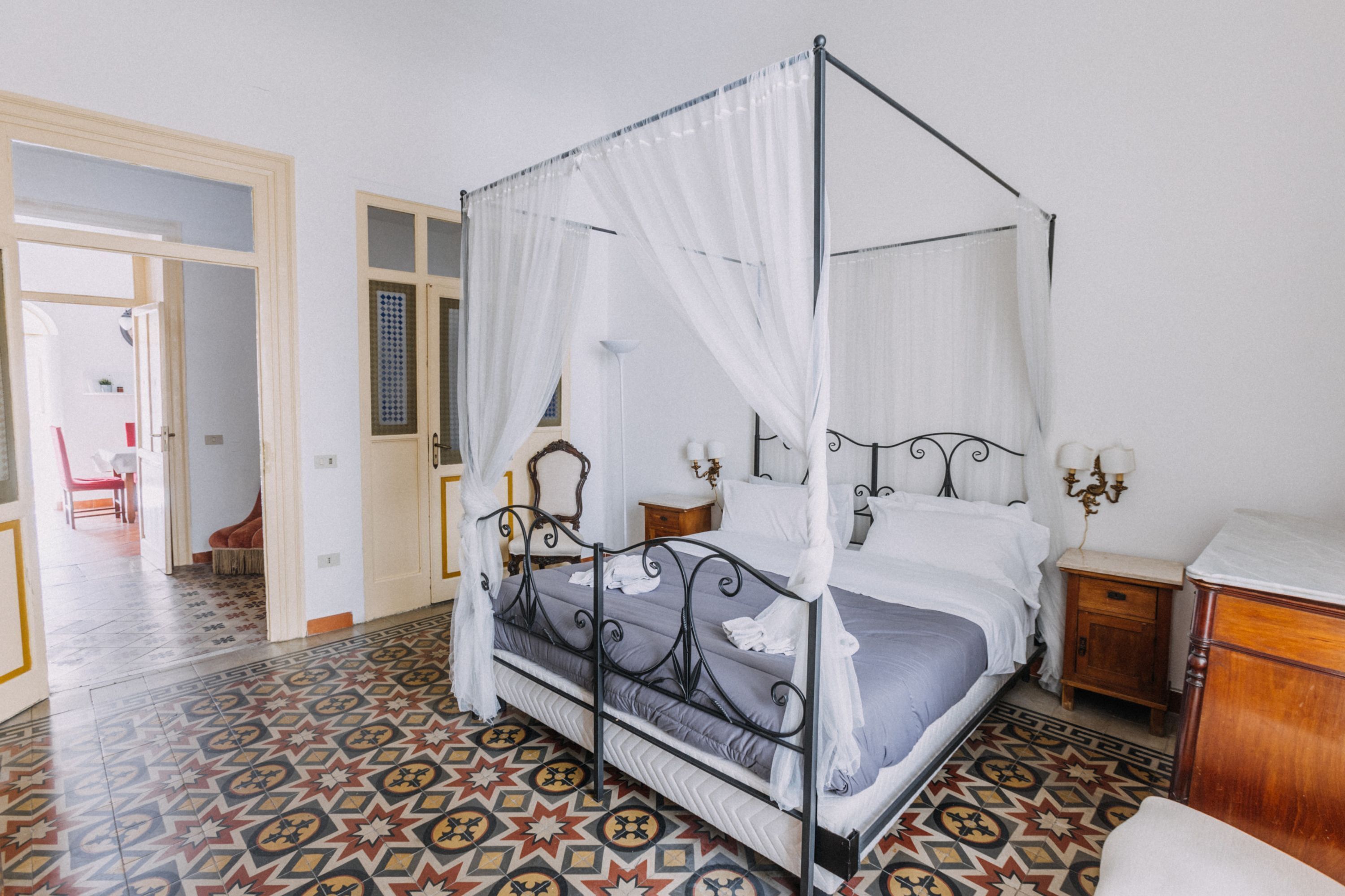 A four-poster bed with a canopy, a bedside table with a lamp, and a patterned floor.