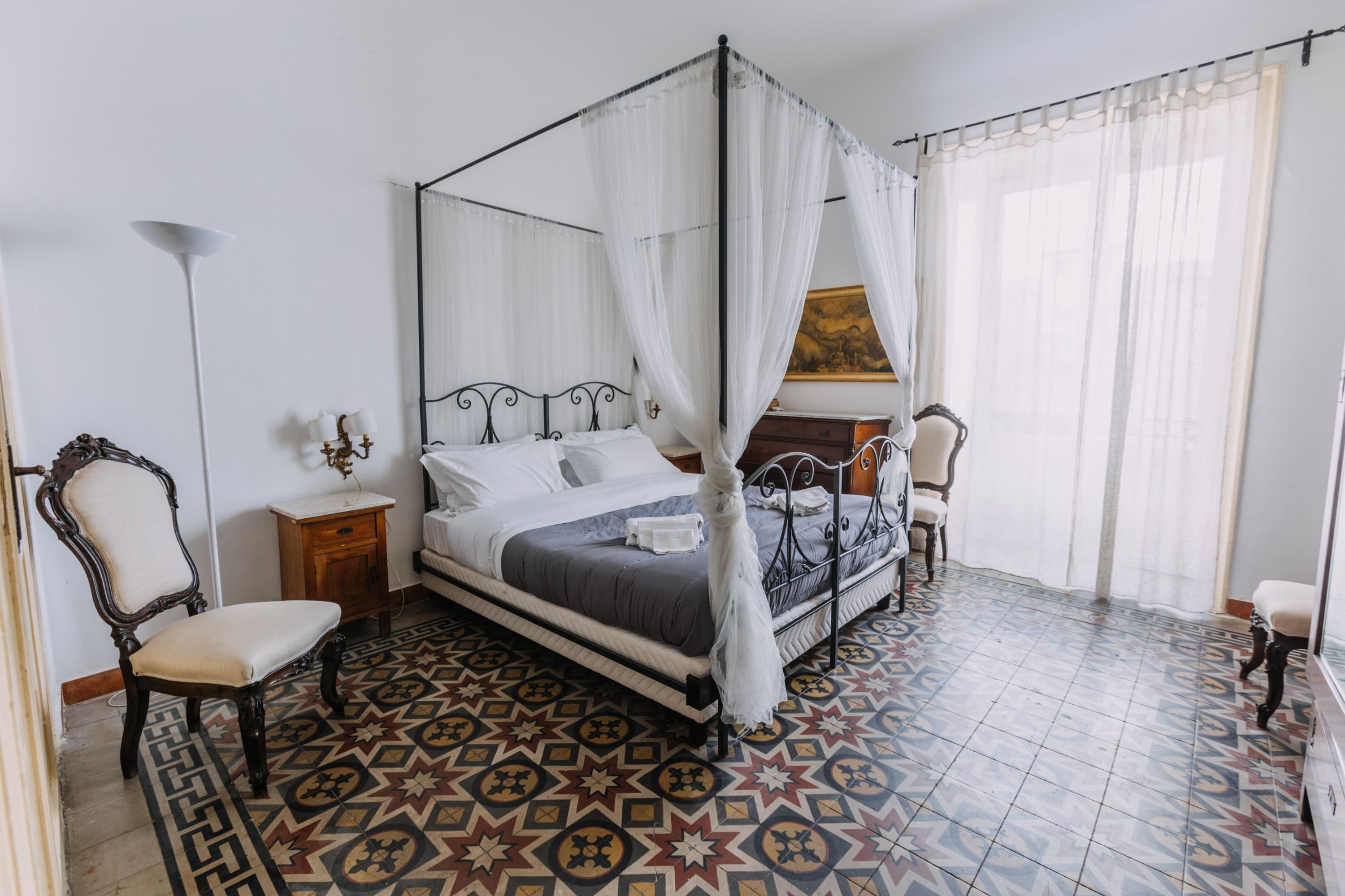 A bedroom with a four-poster bed, a chair, a nightstand, and a floor with patterned tiles.