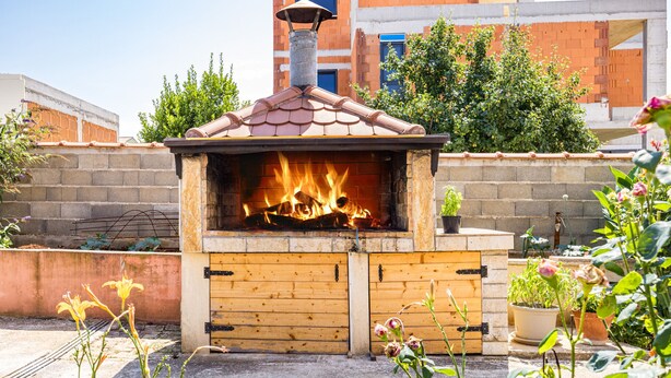 An outdoor brick oven with a tiled roof, surrounded by potted plants and a brick wall.