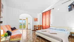 A bedroom with a bed, a sofa, a desk, and a chair. There is a window with curtains, a painting on the wall, and a TV mounted on the wall.