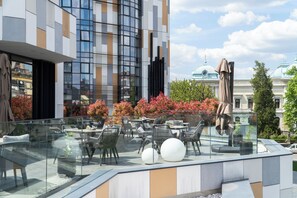 A modern outdoor seating area with glass railings, tables, and chairs, overlooking a cityscape with buildings and trees.