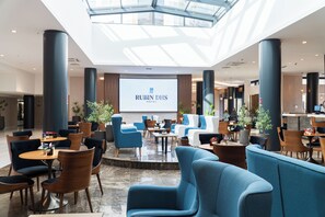 A modern hotel lobby with a large screen displaying the hotel name, comfortable seating areas, and a glass ceiling. - Hotel Rubin DHS (Krusevac)