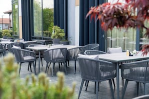 An outdoor seating area with tables and chairs, large windows, and potted plants. - Hotel Rubin DHS (Krusevac)
