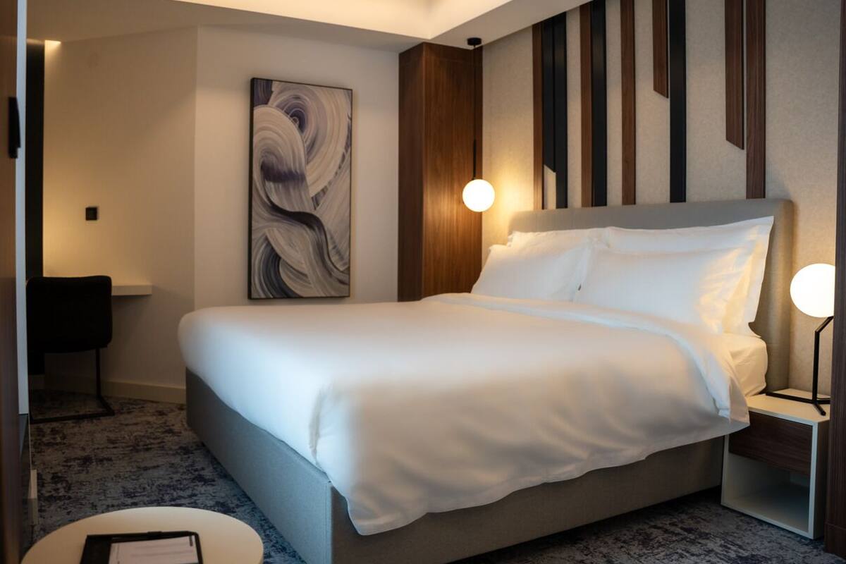A modern hotel room with a large bed, a bedside table, a nightstand, and a wall-mounted lamp.