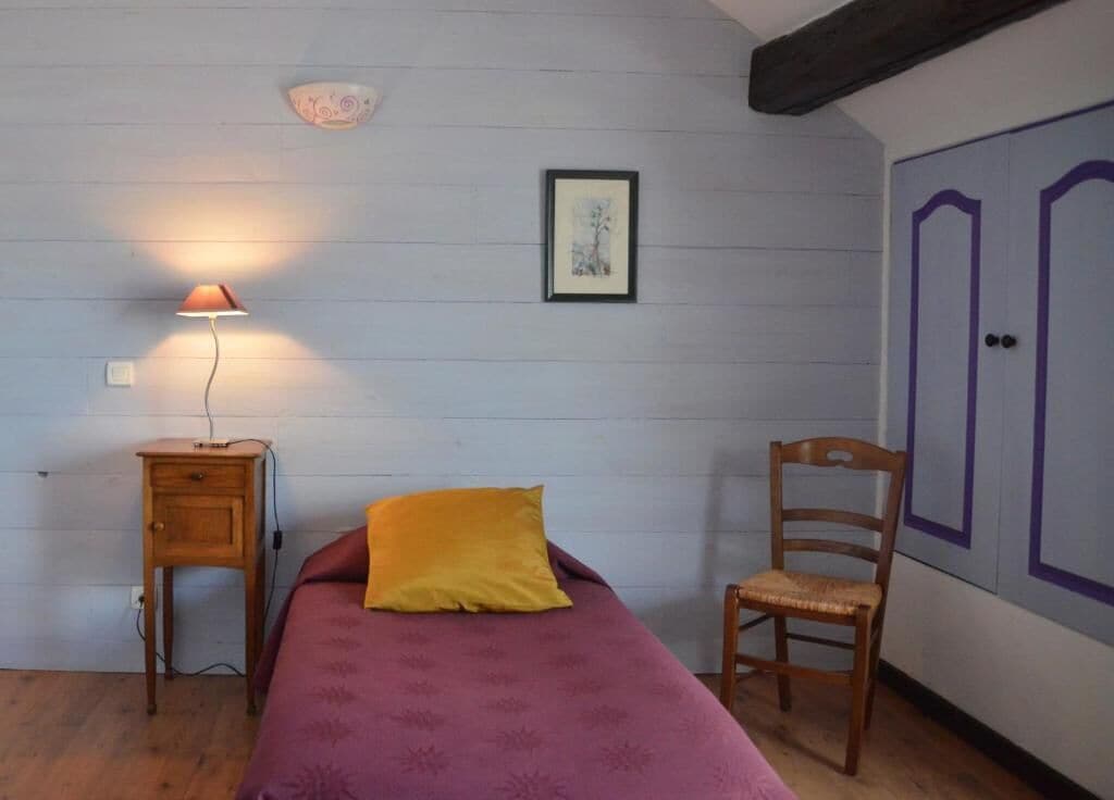 Un dormitorio con cama, mesita de noche con lámpara, una silla, armario y un cuadro en la pared.