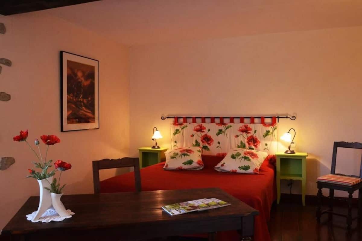 A bedroom with a bed, two bedside tables with lamps, a chair, a table with a magazine, and a vase with flowers.