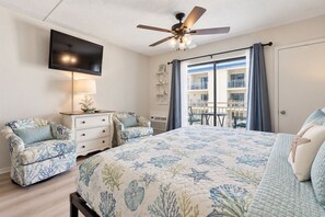 A bedroom with a bed, two chairs, a TV, a ceiling fan, and a balcony with a view.