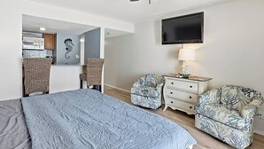 A bedroom with a bed, two chairs, a dresser, a television, and a ceiling fan. - OceanView Studio Condo in Orange Beach | 4 Pools (Orange Beach, AL)