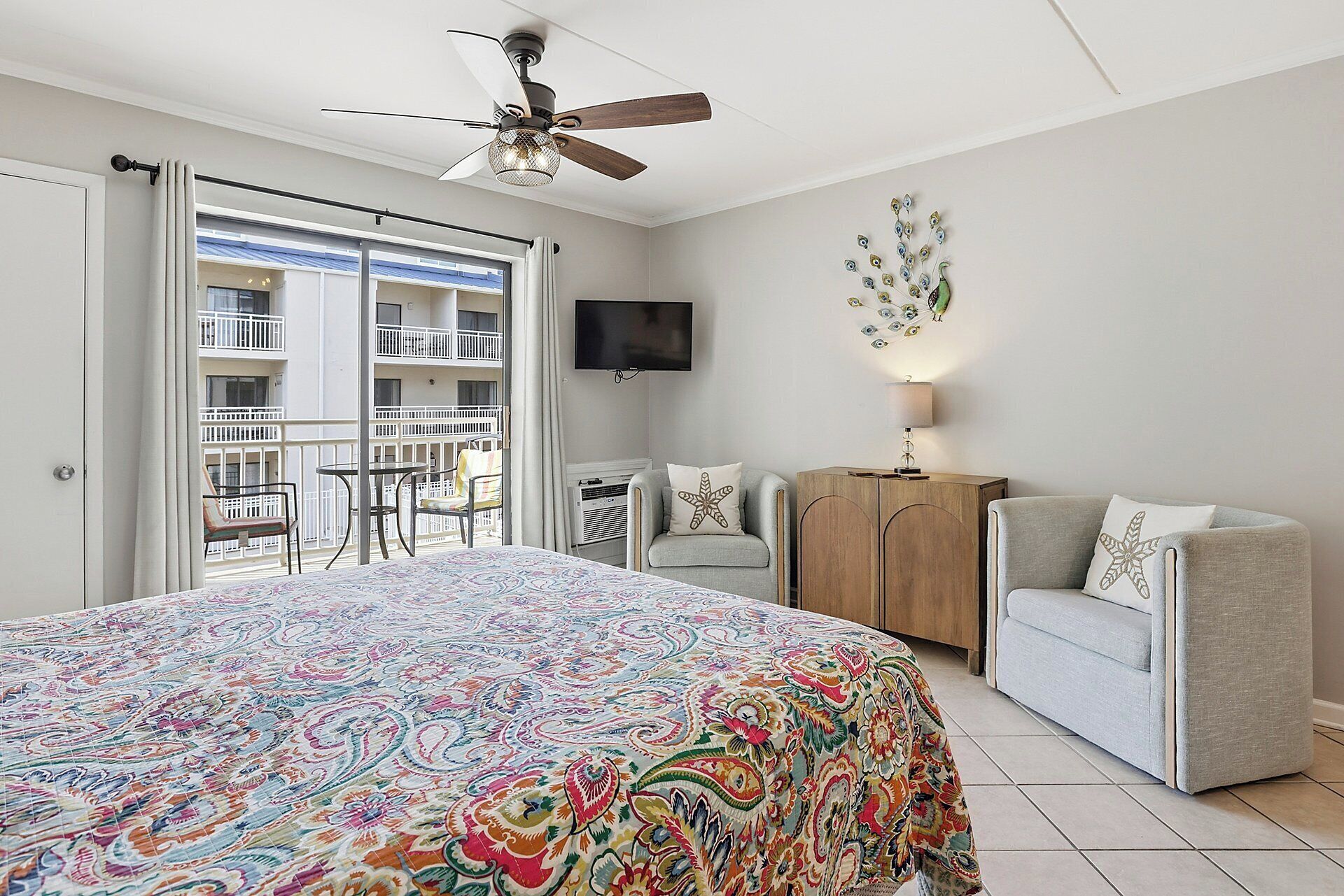 A bedroom with a bed, a ceiling fan, a TV, a balcony with a table and chairs, and a decorative wall piece.