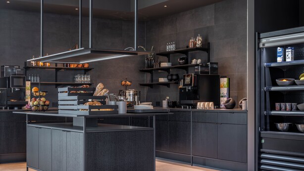 A modern kitchen with dark cabinetry, a central island, and various appliances and utensils. - Zleep Hotel Hamburg Volkspark (Hamburg)