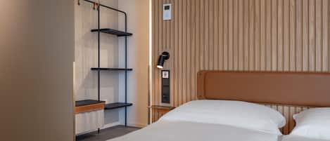 Habitación de hotel con dos camas, cabecero de madera, armario con estantes y mesita de noche con lámpara.