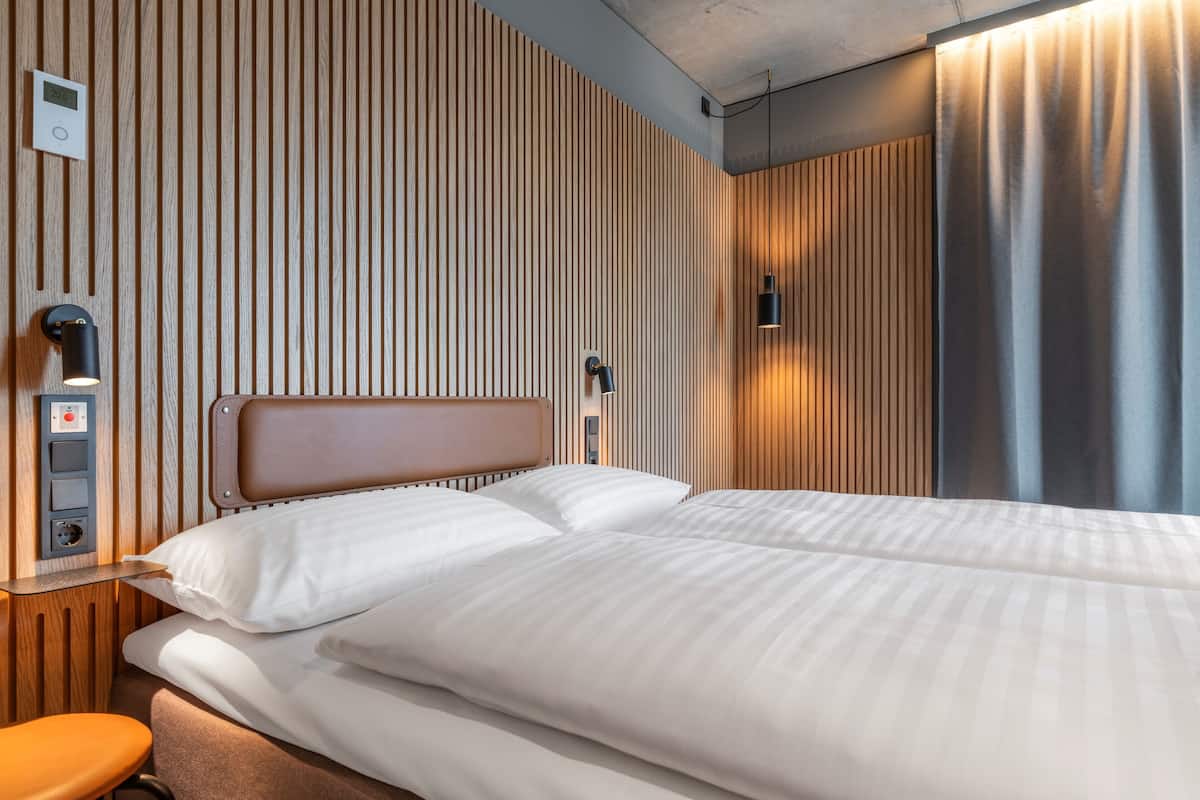 Ein Hotelzimmer mit Bett, Nachttisch, Bedienfeld an der Wand und Vorhang.
