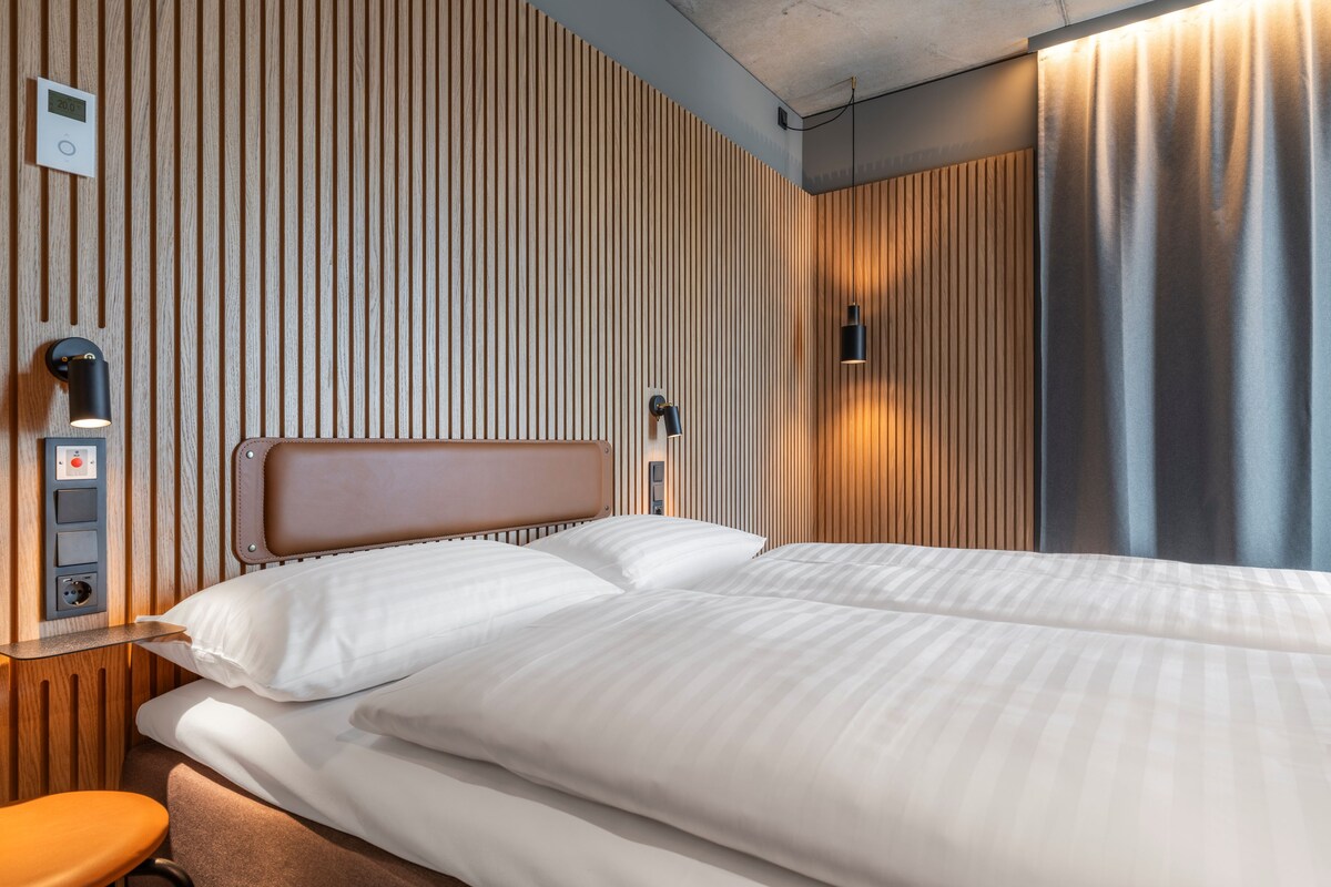 Une chambre d’hôtel avec un lit, une table de chevet, un panneau de commande fixé au mur et un rideau.