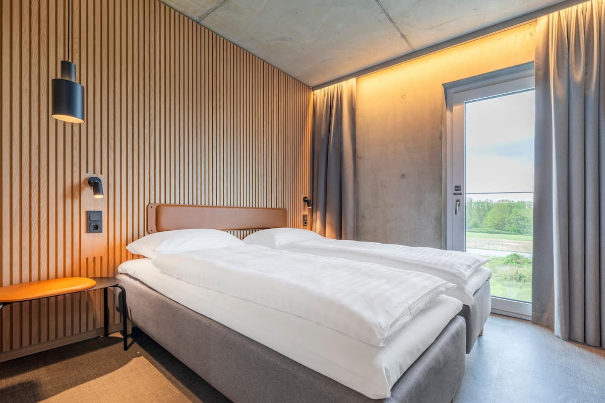 Ein modernes Hotelzimmer mit einem großen Bett, einem Nachttisch, einer Wandlampe und Blick ins Freie durch ein großes Fenster.
