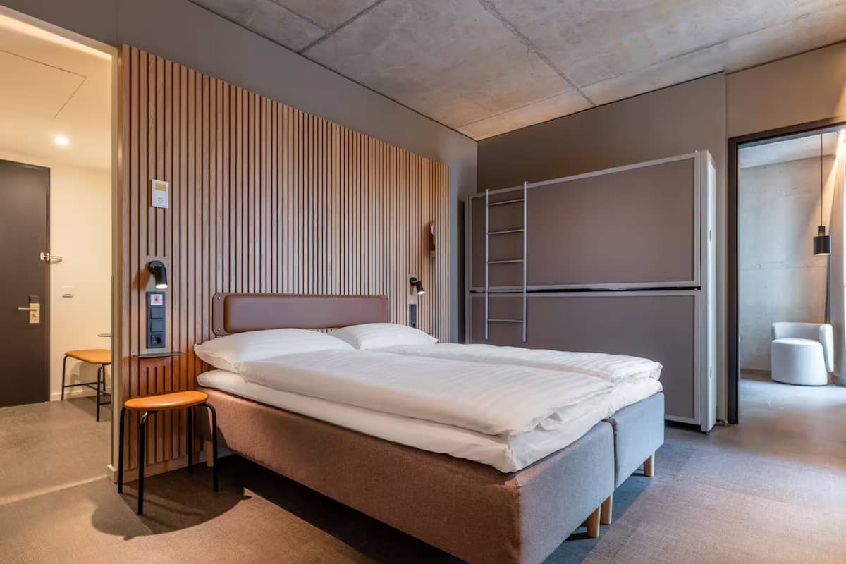 Ein modernes Hotelzimmer mit einem großen Bett, einer Wand aus Holzlamellen, einem kleinen Hocker und einer Leiter.