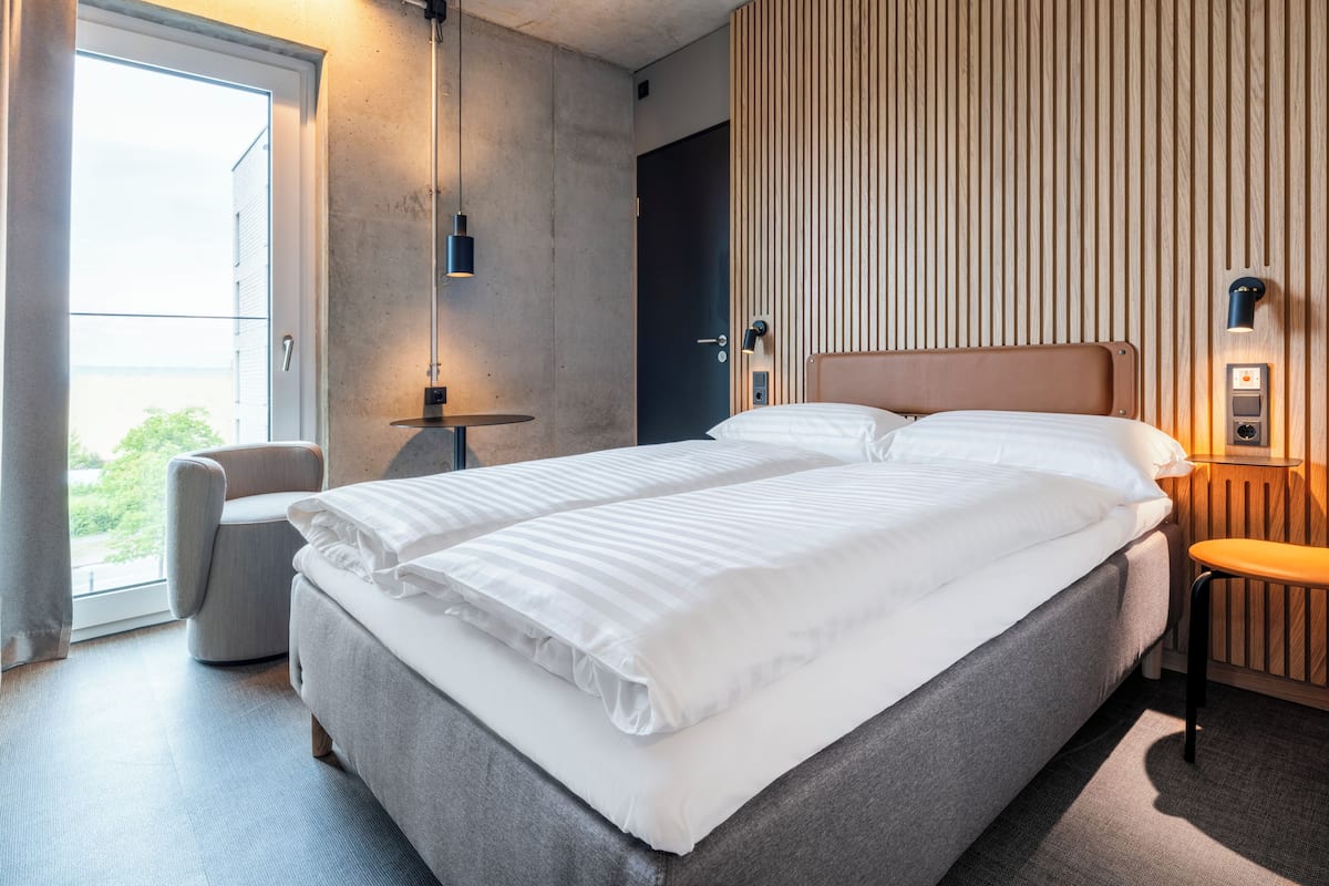 Ein modernes Hotelzimmer mit einem großen Bett, einem kleinen runden Tisch, einem Sessel und Blick ins Freie.