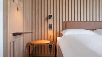 A hotel room with a bed, a small table, and a wall-mounted shelf.