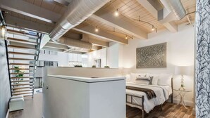 Quarto de hotel moderno com uma cama grande, uma mesinha de cabeceira pequena, um detalhe decorativo na parede e uma escada para o andar superior.