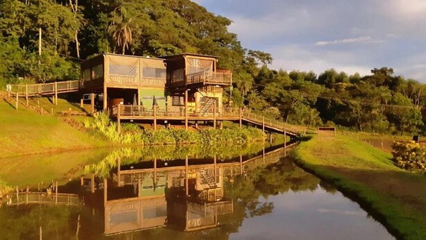 Una casa de madera con balcón, ubicada junto a una masa de agua tranquila, rodeada de vegetación.