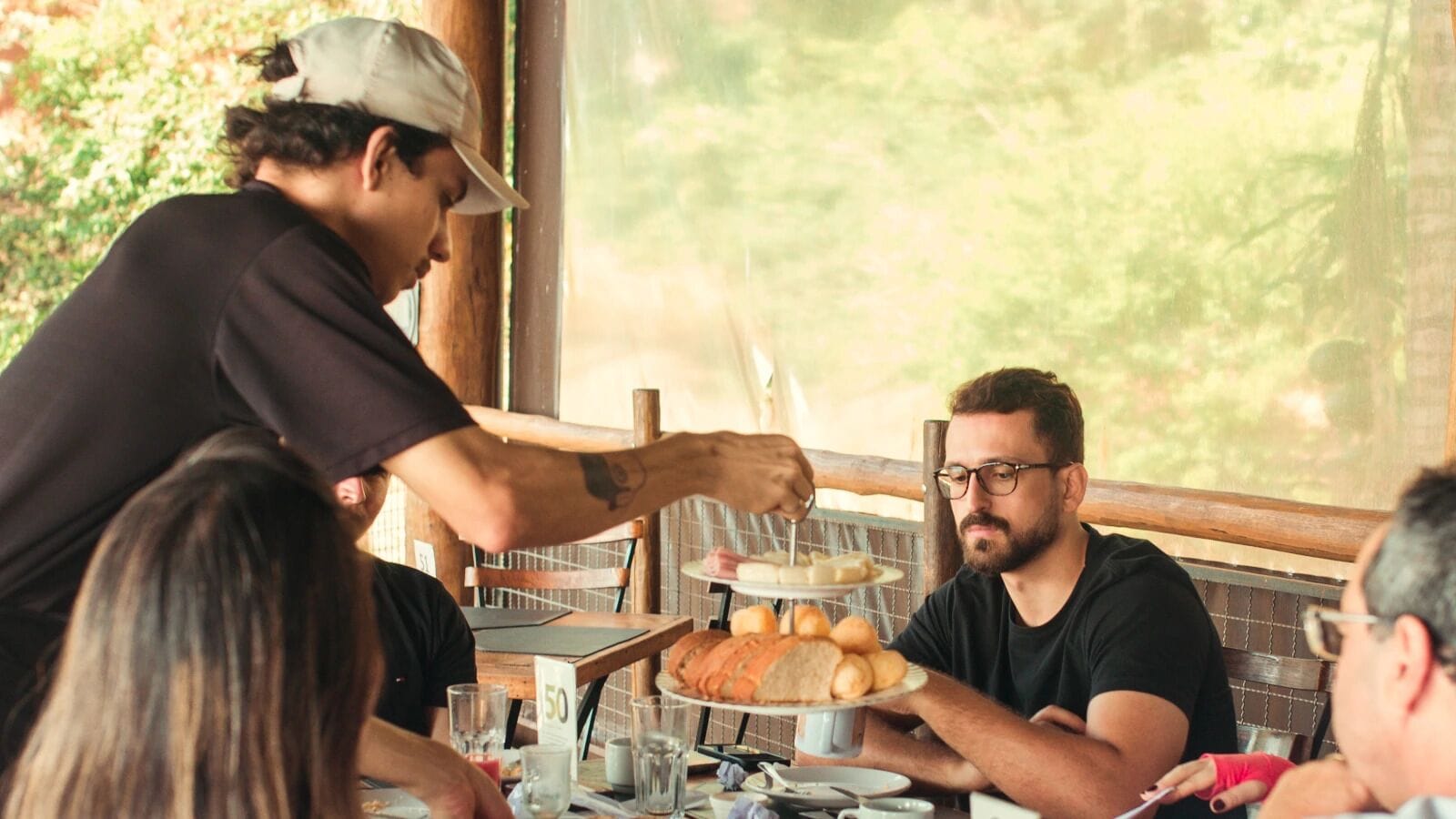 A person serving food to a group at a table with a view of greenery outside.