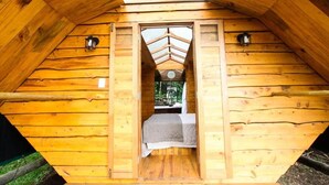 Una cabaña de madera con techo a dos aguas, una cama con una manta gris y dos faroles fijados a la pared.