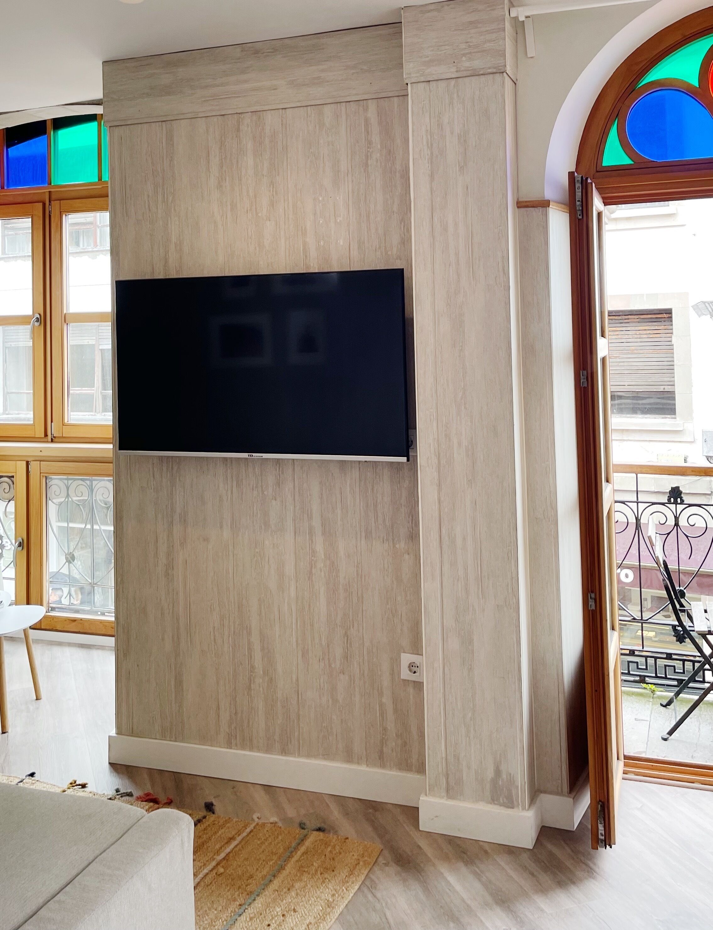 A room with a large flat-screen TV mounted on a wooden wall, a window with a view of the outside, and a door leading to an outdoor area.