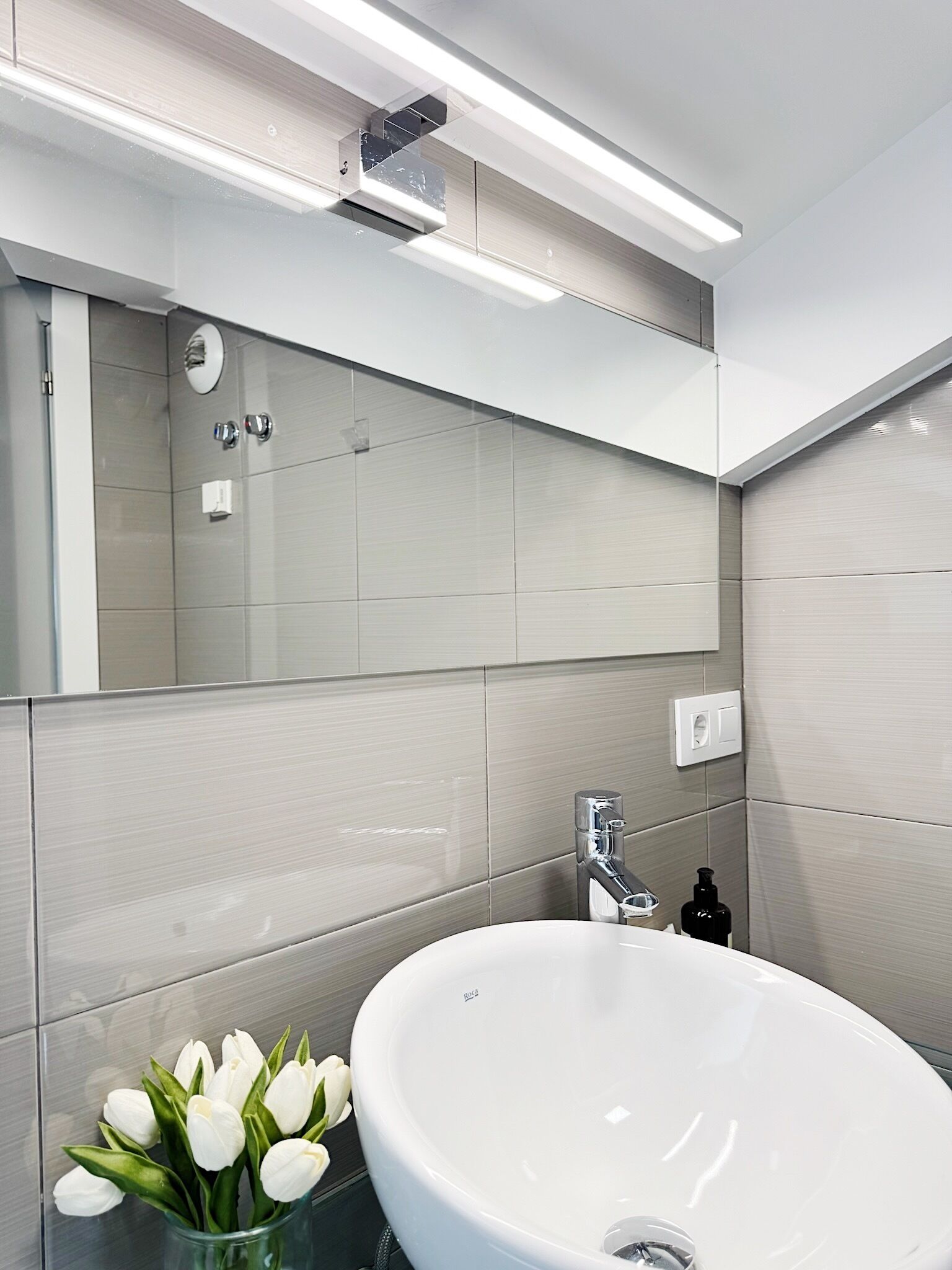 Un baño moderno con un lavamanos grande de color blanco, un espejo y un florero con flores blancas.