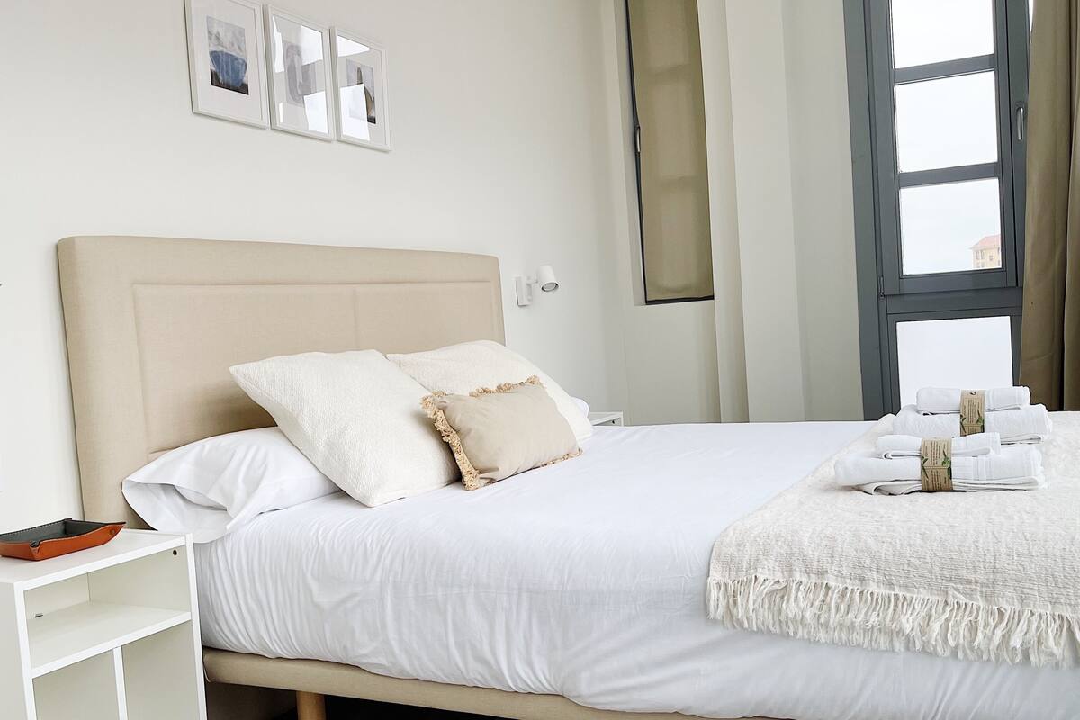 Una cama bien hecha con almohadas, una mesita de noche con una bandeja y una ventana con cortinas.