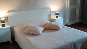 Quarto com cama branca, duas mesas de cabeceira com abajures, piso de madeira e uma porta ao fundo.