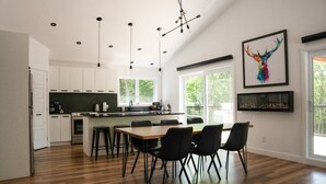 Cocina moderna con armarios blancos, una mesa de comedor de madera y sillas negras. Hay luces colgantes y un ventanal grande que proporciona luz natural.
