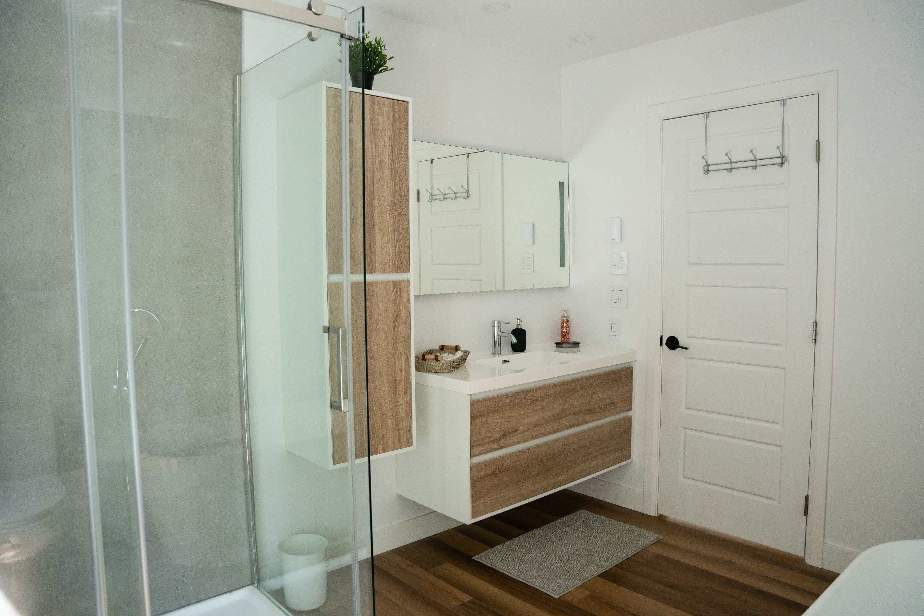 Un baño moderno con cabina de ducha de vidrio, tocador de madera y una puerta blanca con perchero.