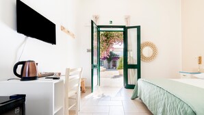 A hotel room with a bed, a desk with a kettle, a chair, a TV mounted on the wall, and an open door leading to a balcony with plants.