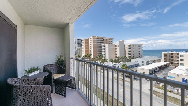Ein Balkon mit Korbmöbeln, Blick auf einen Strand mit Gebäuden und einem klaren Himmel.