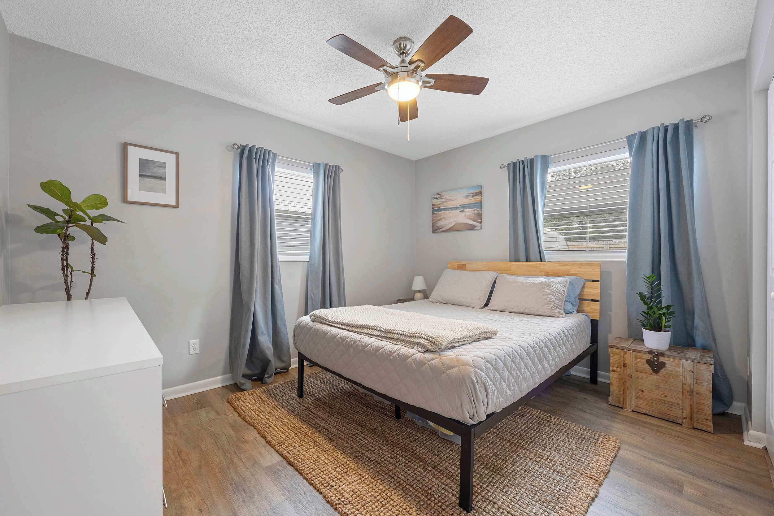 A bedroom with a bed, a ceiling fan, a nightstand, a potted plant, and a window with curtains.