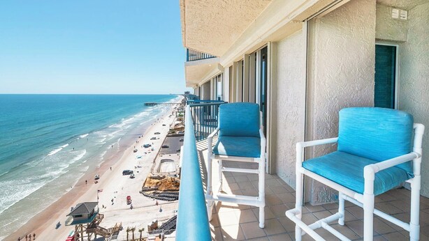 A balcony with two blue cushioned chairs, overlooking a beach with people and umbrellas.