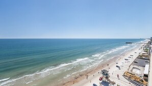 A beach with a pier extending into the ocean, a sandy shoreline with people, and a parking area adjacent to the beach.