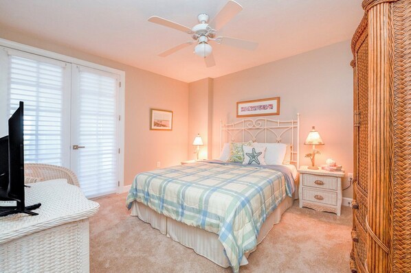 A bedroom with a bed, two bedside tables with lamps, a ceiling fan, a framed picture on the wall, and a wicker chair.