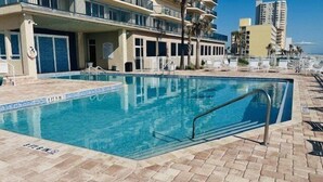 A swimming pool with a tiled deck, lounge chairs, and a building with balconies in the background.