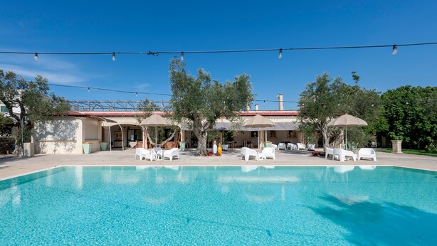 A clear swimming pool with lounge chairs and umbrellas, surrounded by trees and a building with a red roof. - Room 'San Biagio - Superior 105' with Shared Pool, Wi-Fi & Air Conditioning (Calimera)