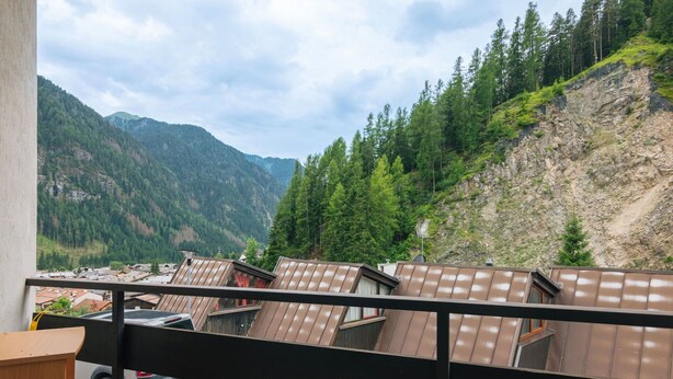 Un balcone con vista su un paesaggio montuoso, che include un pendio boscoso e una valle con edifici.