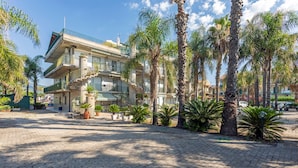 A multi-story building with balconies, palm trees, and a paved courtyard. - Apartment 'I Ciottoli Dello Ionio - Apt 1' with Mountain View, Shared Terrace and Balcony (Mascali)