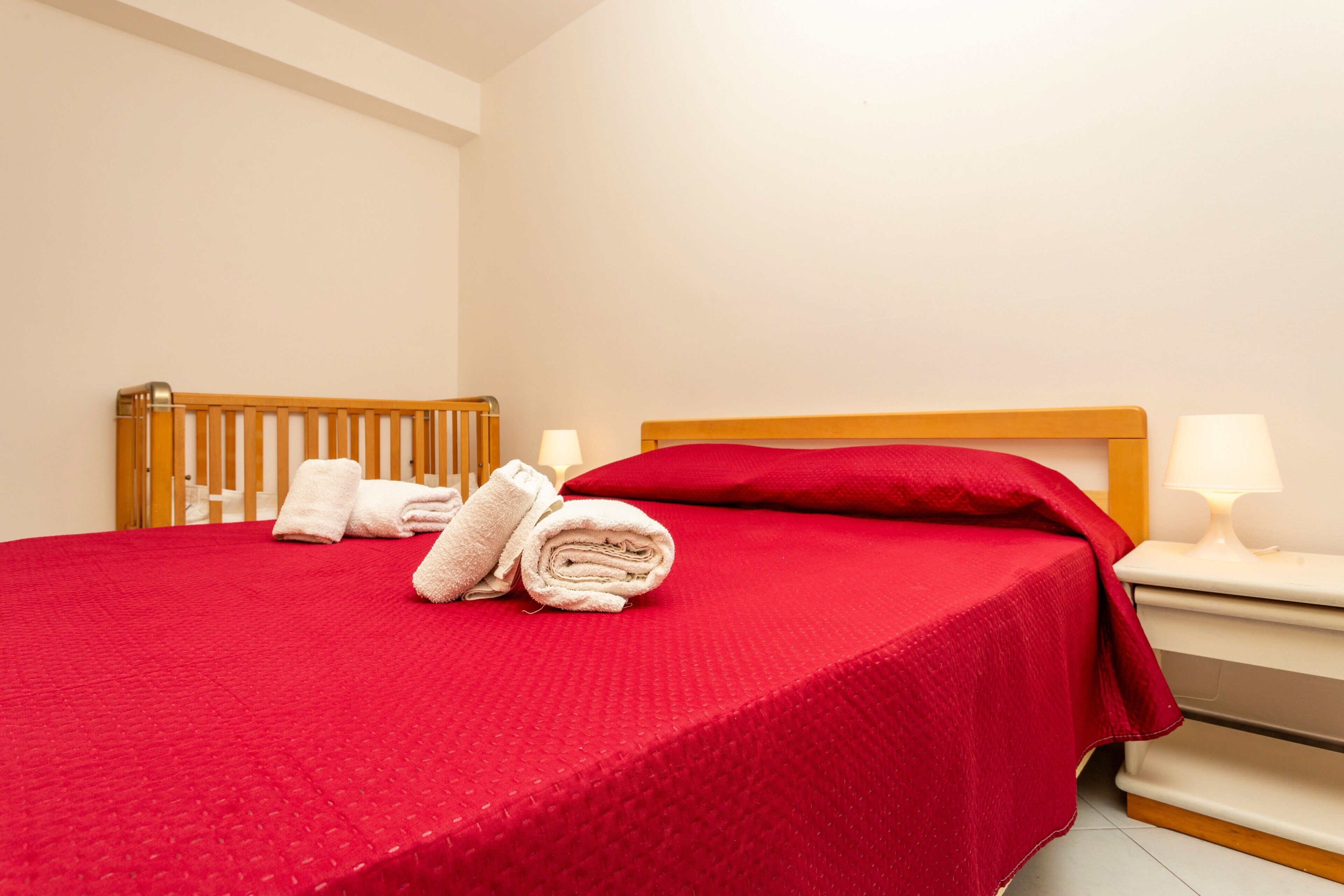 A neatly made bed with a red bedspread, a wooden headboard, and two rolled towels.