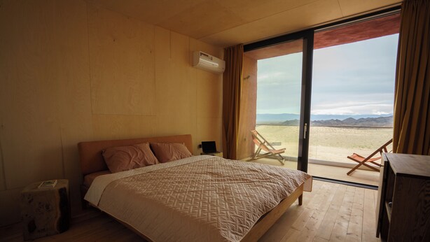 A bedroom with a large bed, a wooden floor, a window with a view of a desert landscape, and an air conditioning unit. -  Pana Unique Charyn (Charyn Canyon)
