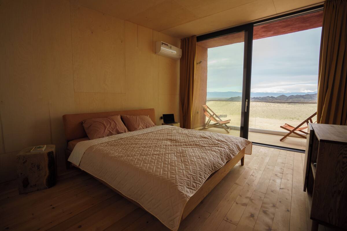 A bedroom with a large bed, a wooden floor, a window with a view of a desert landscape, and an air conditioning unit.